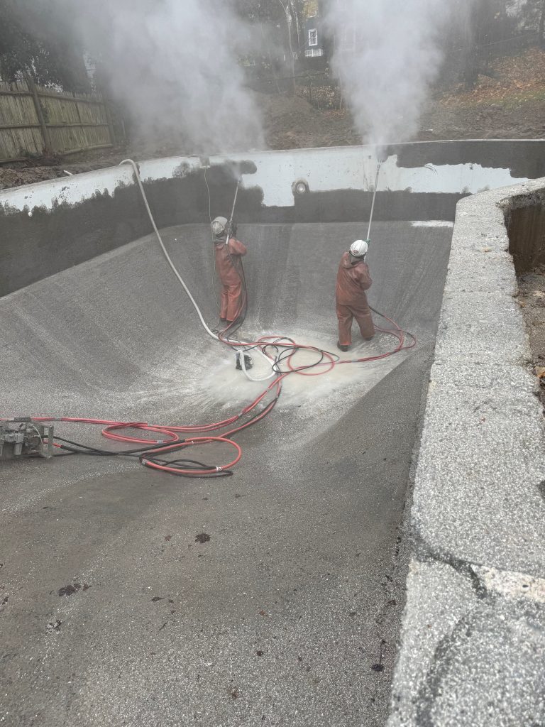 Two men in an inground pool, using HP water hand lances to remove a pool coating.
