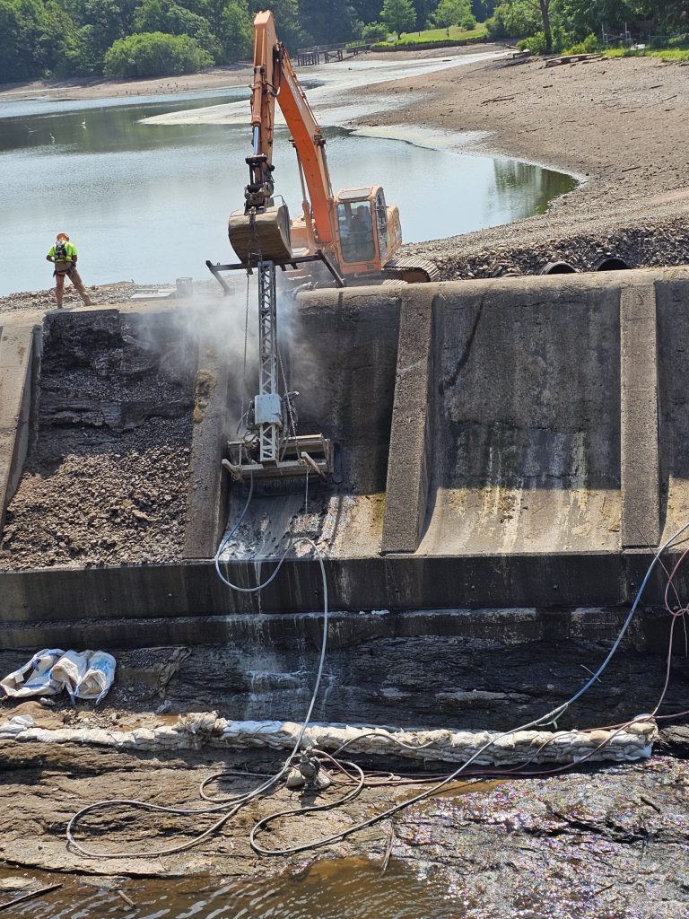 An operator watching the robot head attached to an excavator as it performs hydrodemolition on the dam face.
